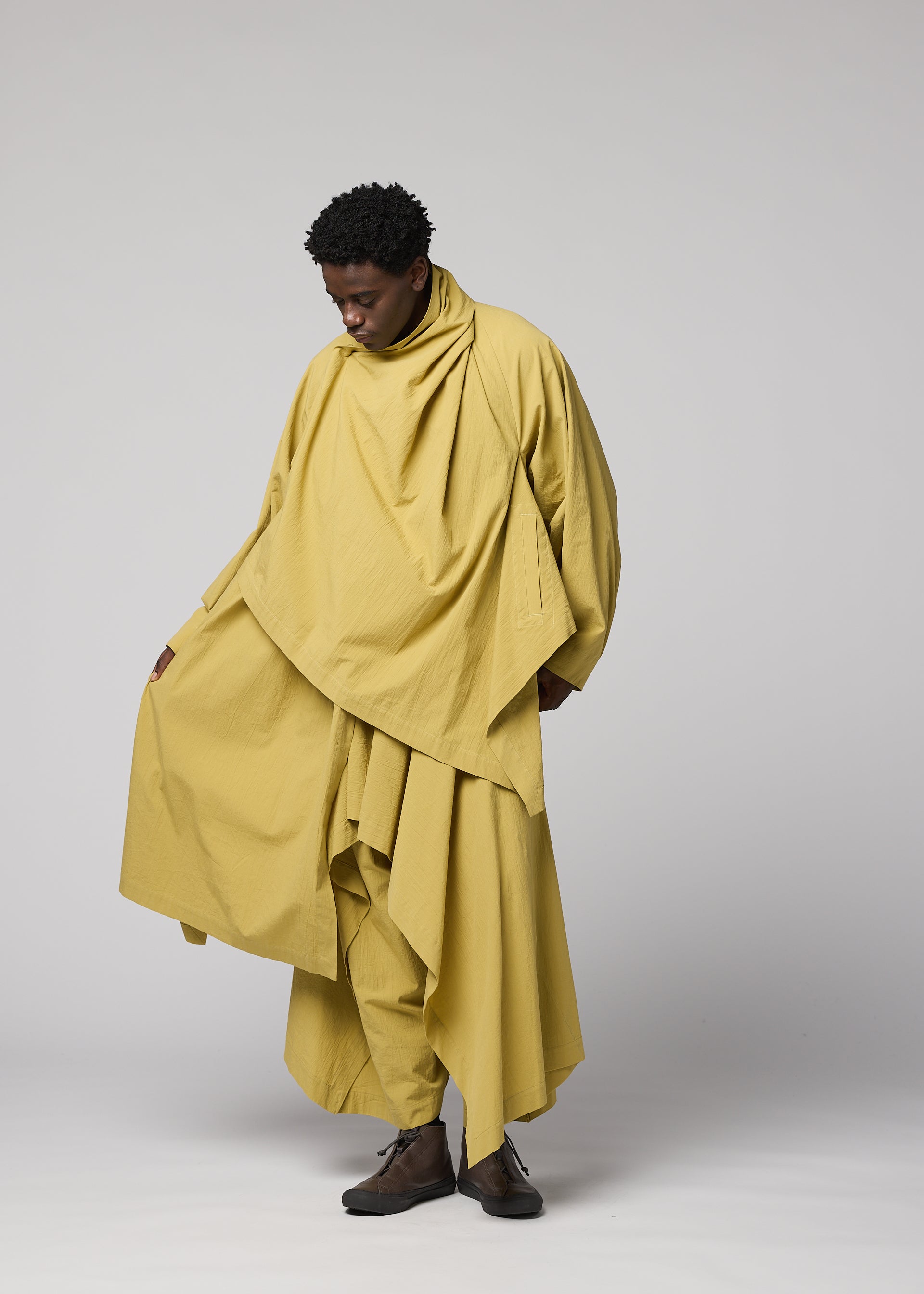 Zoomed view Against a plain backdrop, a person wears loose, layered, oversized yellow clothing from IM MEN’s COMFORT COTTON collection, featuring an irregular-cut jacket made of high-twist organic cotton. With dark shoes and one hand holding the garment, they embody versatile styling while gazing downward thoughtfully.