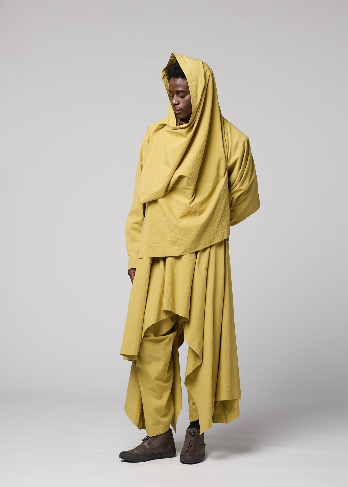 Against a plain backdrop, a person models the COMFORT COTTON ensemble by IM MEN—a flowing, layered outfit in yellow made from high-twist organic cotton and designed for versatile styling. The look includes a hooded scarf, loose top, wide pants, and an irregular-cut jacket. Posing with one hand behind their back, they gaze to the side. view 9 of 9