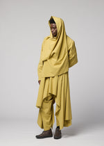 Against a plain backdrop, a person models the COMFORT COTTON ensemble by IM MEN—a flowing, layered outfit in yellow made from high-twist organic cotton and designed for versatile styling. The look includes a hooded scarf, loose top, wide pants, and an irregular-cut jacket. Posing with one hand behind their back, they gaze to the side.