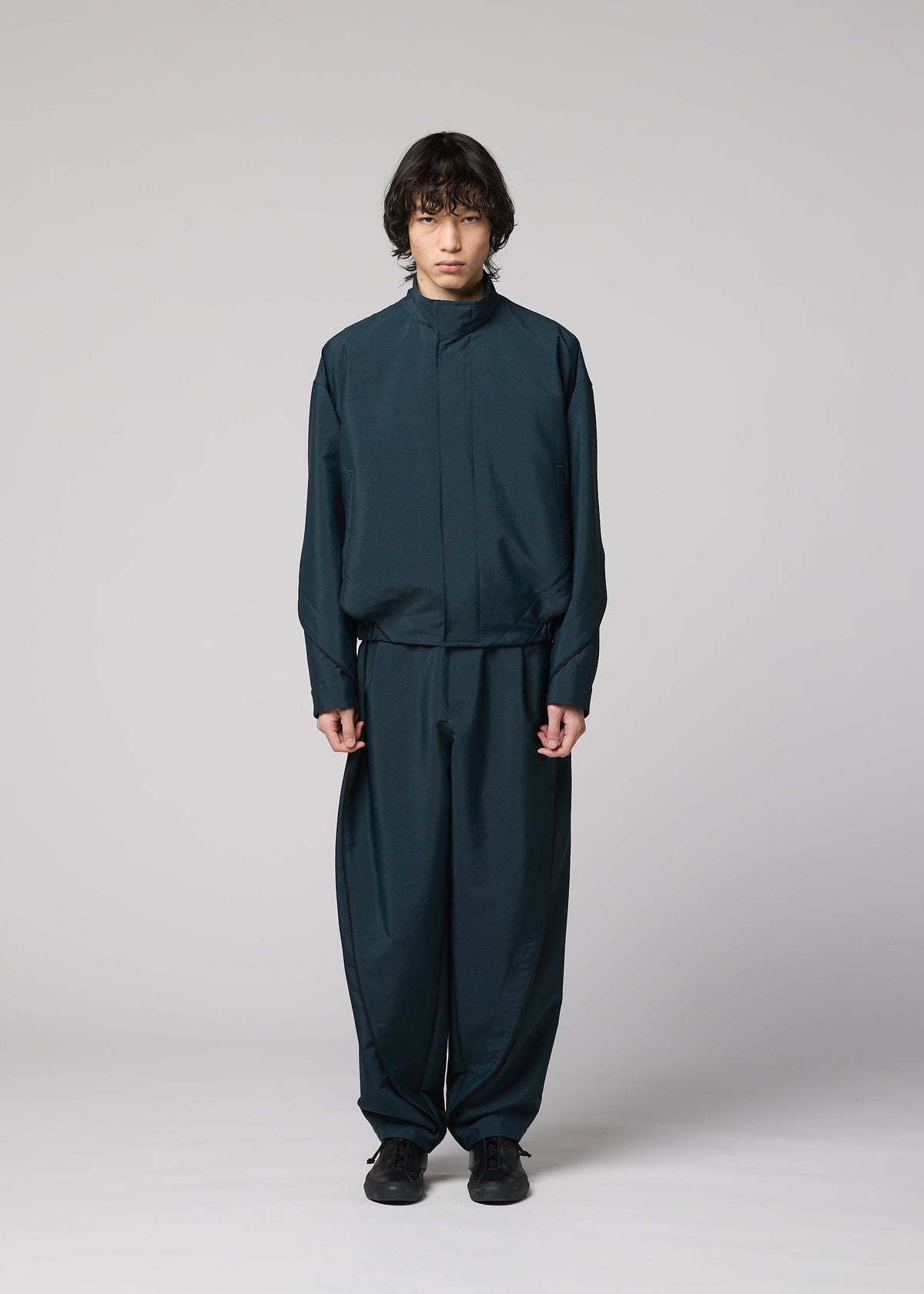 A person with medium-length dark hair faces forward, wearing the IM MEN FLUENT wrinkle-resistant dark teal jacket and matching wide-leg pants, paired with black shoes, against a plain light gray background. view 6 of 9