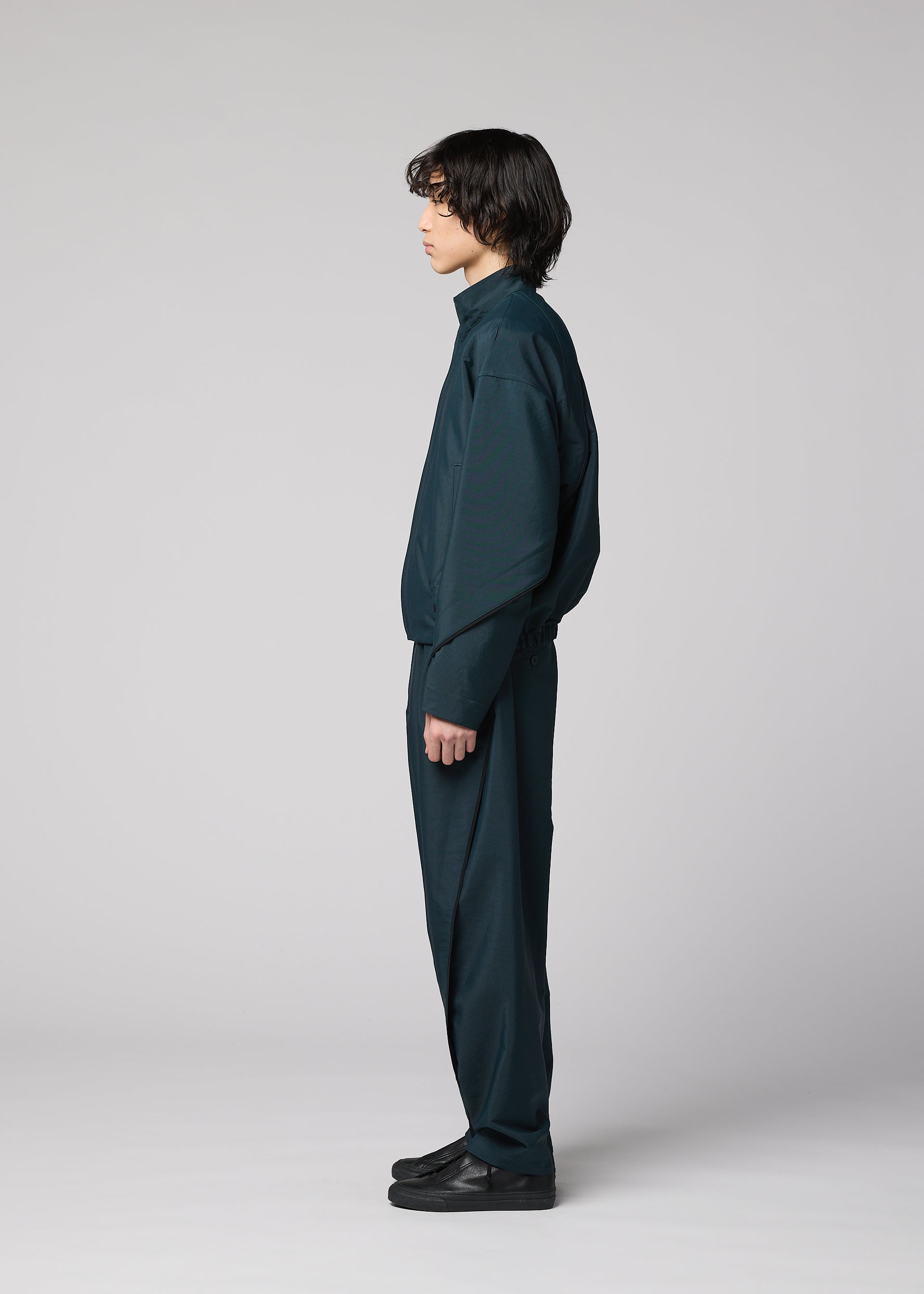 Zoomed view A person with medium-length dark hair stands in profile against a light gray background, wearing the IM MEN FLUENT dark green wrinkle-resistant long-sleeve shirt and pants, paired with black shoes.