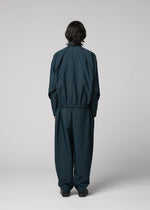 A person with medium-length dark hair faces away from the camera, wearing the IM MEN FLUENT lightweight loose-fitting jacket and matching pants in dark teal, set against a plain light gray background.