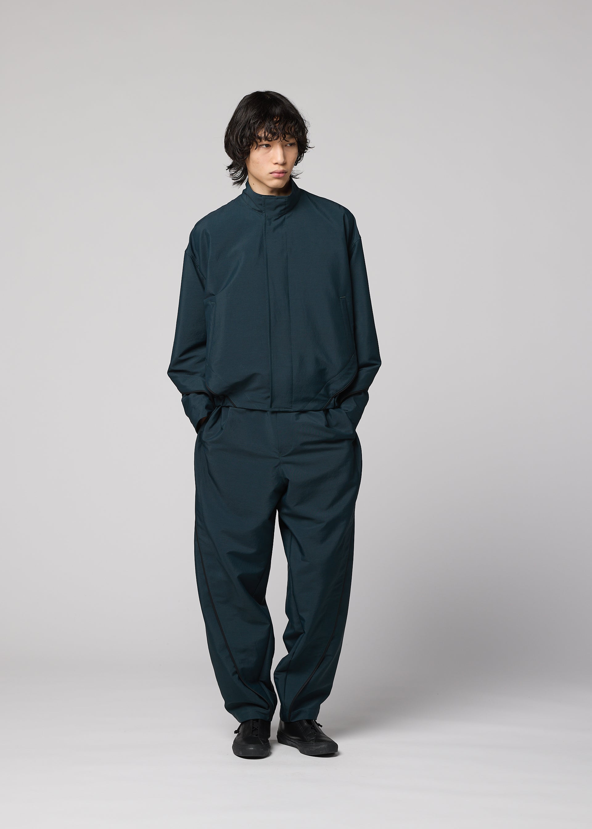 Zoomed view A person with medium-length dark hair stands against a light gray background, wearing the IM MEN FLUENT wrinkle-resistant set—dark teal, loose-fitting jacket and matching pants with long sleeves and pockets—paired with black shoes.