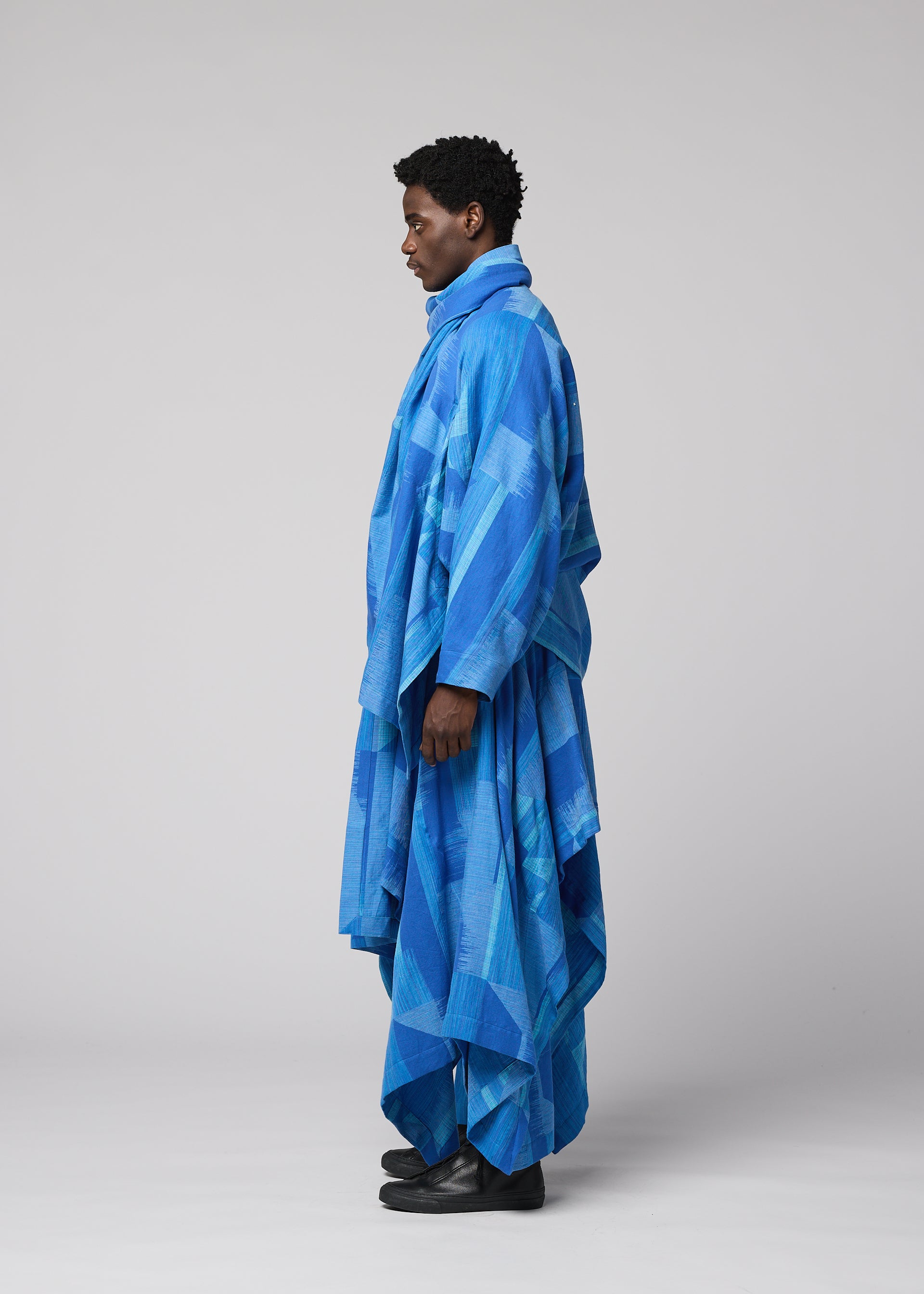Zoomed view A person stands in profile wearing the IM MEN KASURI, a long, loose-fitting blue jacket with traditional Kasuri weaving patterns, paired with black shoes against a plain light gray background.