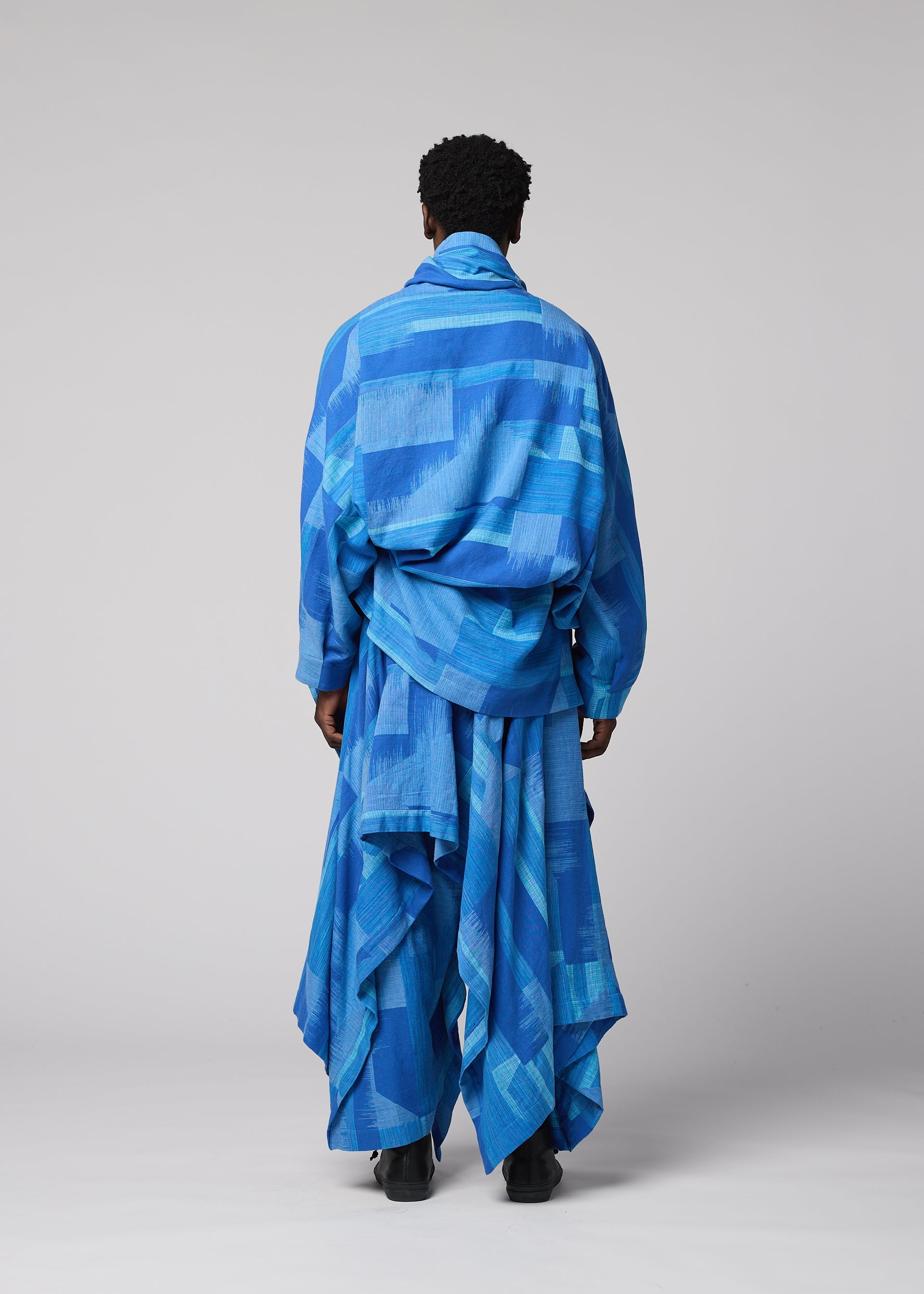 Zoomed view Someone stands with their back to the camera, dressed in IM MEN’s KASURI set—a relaxed, draped blue ensemble featuring geometric patterns inspired by traditional Kasuri weaving. The background is a plain light gray.
