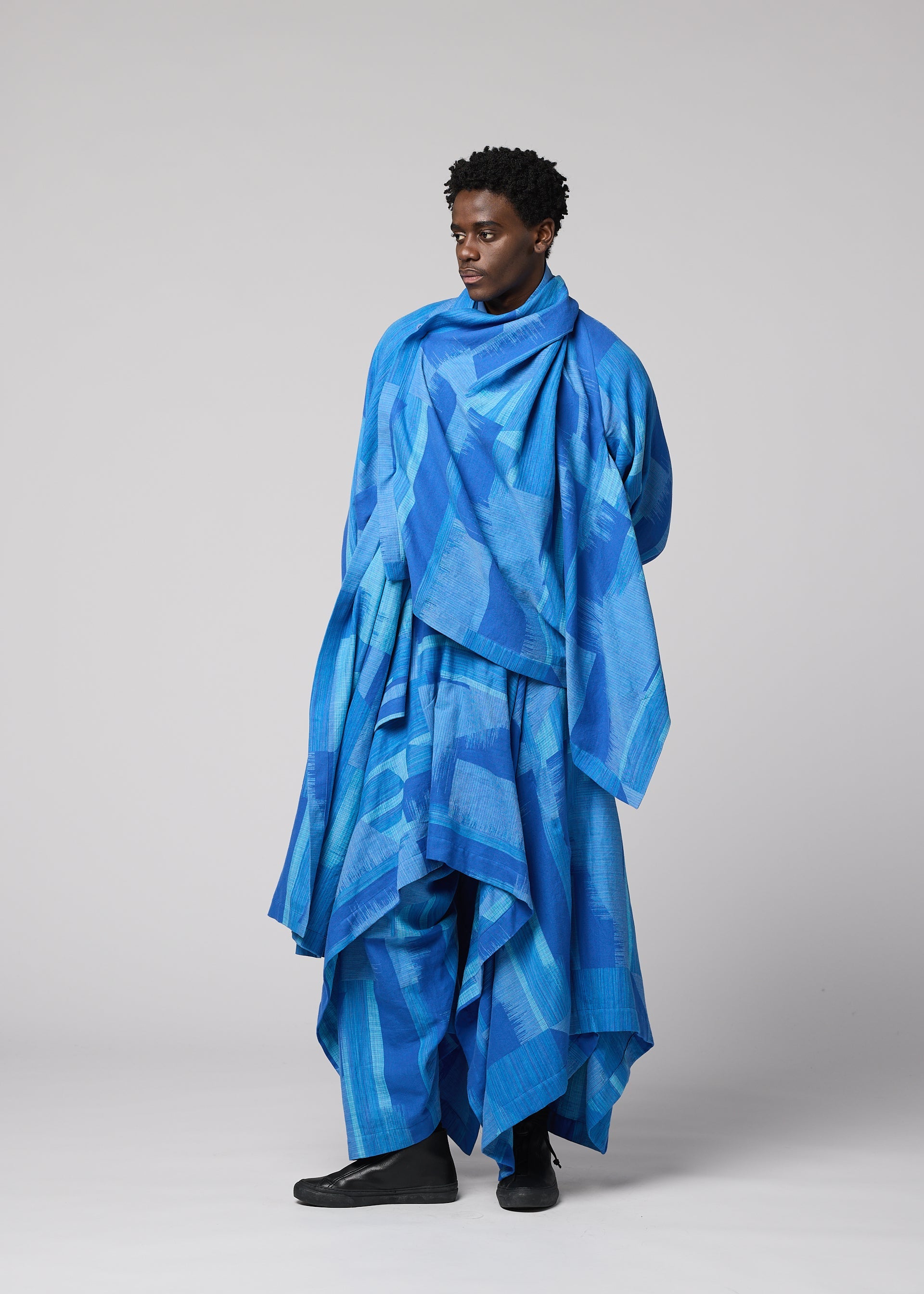 Zoomed view A person stands wearing an oversized, layered outfit in various shades of blue with abstract patterns, featuring the distinctive IM MEN KASURI jacket draped elegantly, looking to the side against a plain light gray background.