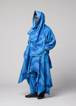 A person stands against a plain background, wearing IM MEN’s KASURI—a layered, flowing blue drape jacket featuring geometric patterns inspired by Kasuri weaving and a hood, covering most of their body except for the face and hands.