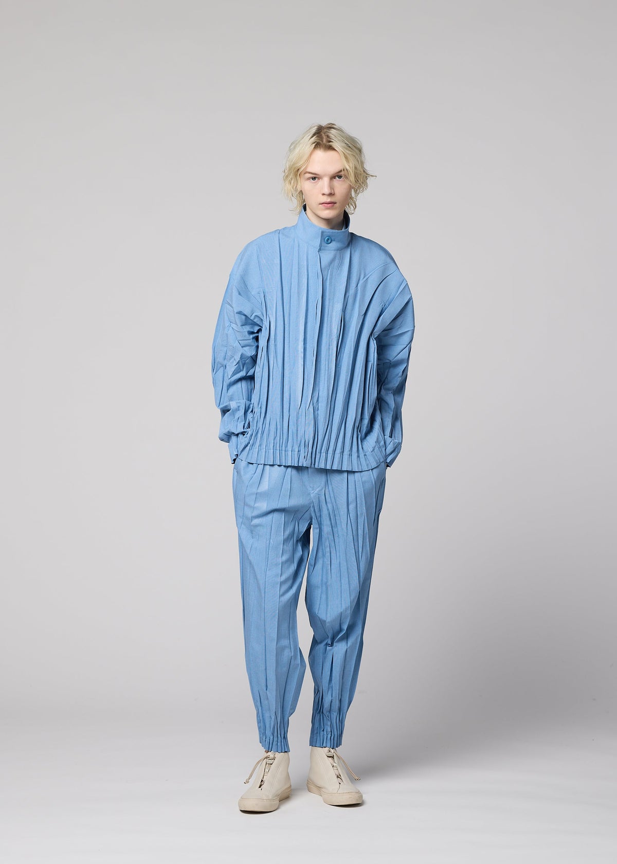 A person with blonde hair wears the IM MEN EDGE ensemble—a light blue, pleated jacket made from mélange polyester paired with matching pants and cream-colored shoes—standing against a plain gray background. The three-dimensional design of EDGE lends remarkable depth and sophistication to the overall style. view 9 of 9