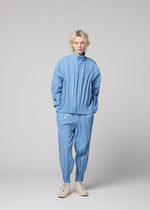 A person with blonde hair wears the IM MEN EDGE ensemble—a light blue, pleated jacket made from mélange polyester paired with matching pants and cream-colored shoes—standing against a plain gray background. The three-dimensional design of EDGE lends remarkable depth and sophistication to the overall style.
