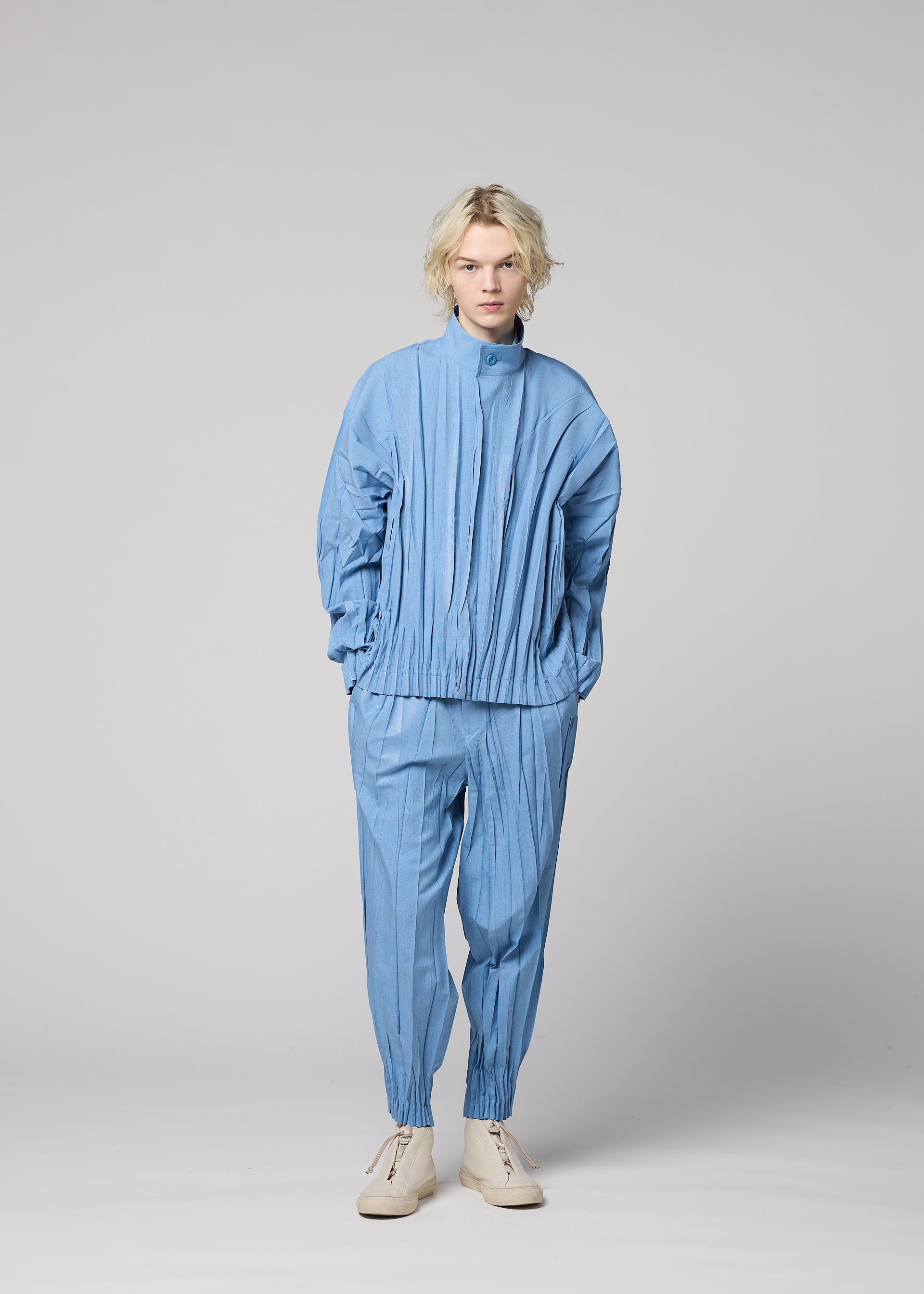 Zoomed view A person with blonde hair wears the IM MEN EDGE ensemble—a light blue, pleated jacket made from mélange polyester paired with matching pants and cream-colored shoes—standing against a plain gray background. The three-dimensional design of EDGE lends remarkable depth and sophistication to the overall style.