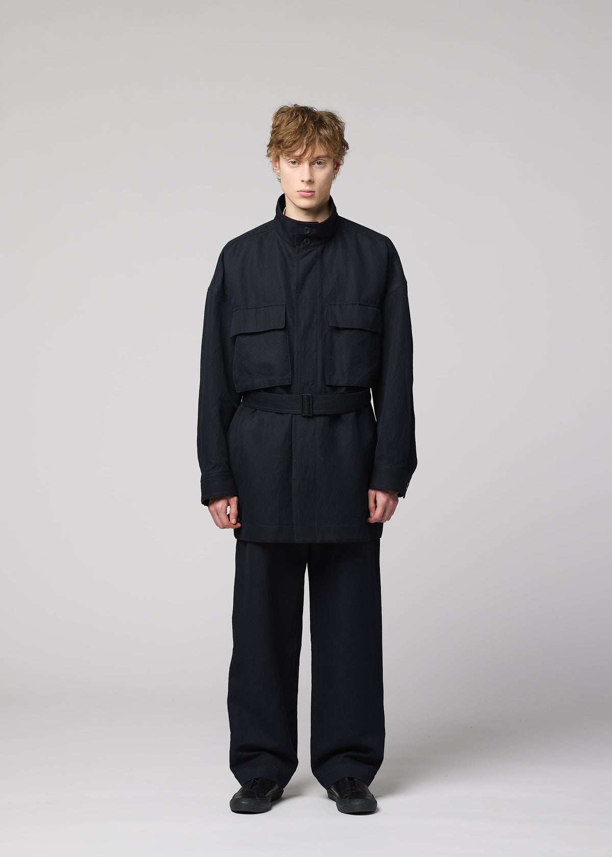 A person with short, curly hair stands in a studio wearing the IM MEN TOUGH WOOL COTTON black belted jacket made from luxurious Bishu wool, featuring large front pockets, paired with black pants and black shoes against a plain light gray background. view 6 of 9
