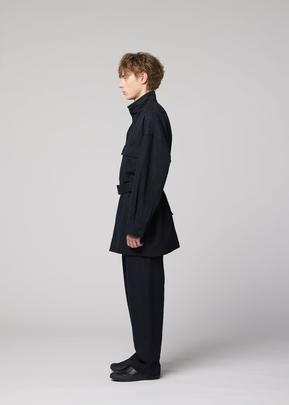 A person stands in profile against a plain gray background, dressed in the IM MEN TOUGH WOOL COTTON jacket and matching black pants, paired with black shoes. The sleek ensemble features luxurious Bishu wool blend fabric, and the individual’s posture is upright, facing left. view 7 of 9