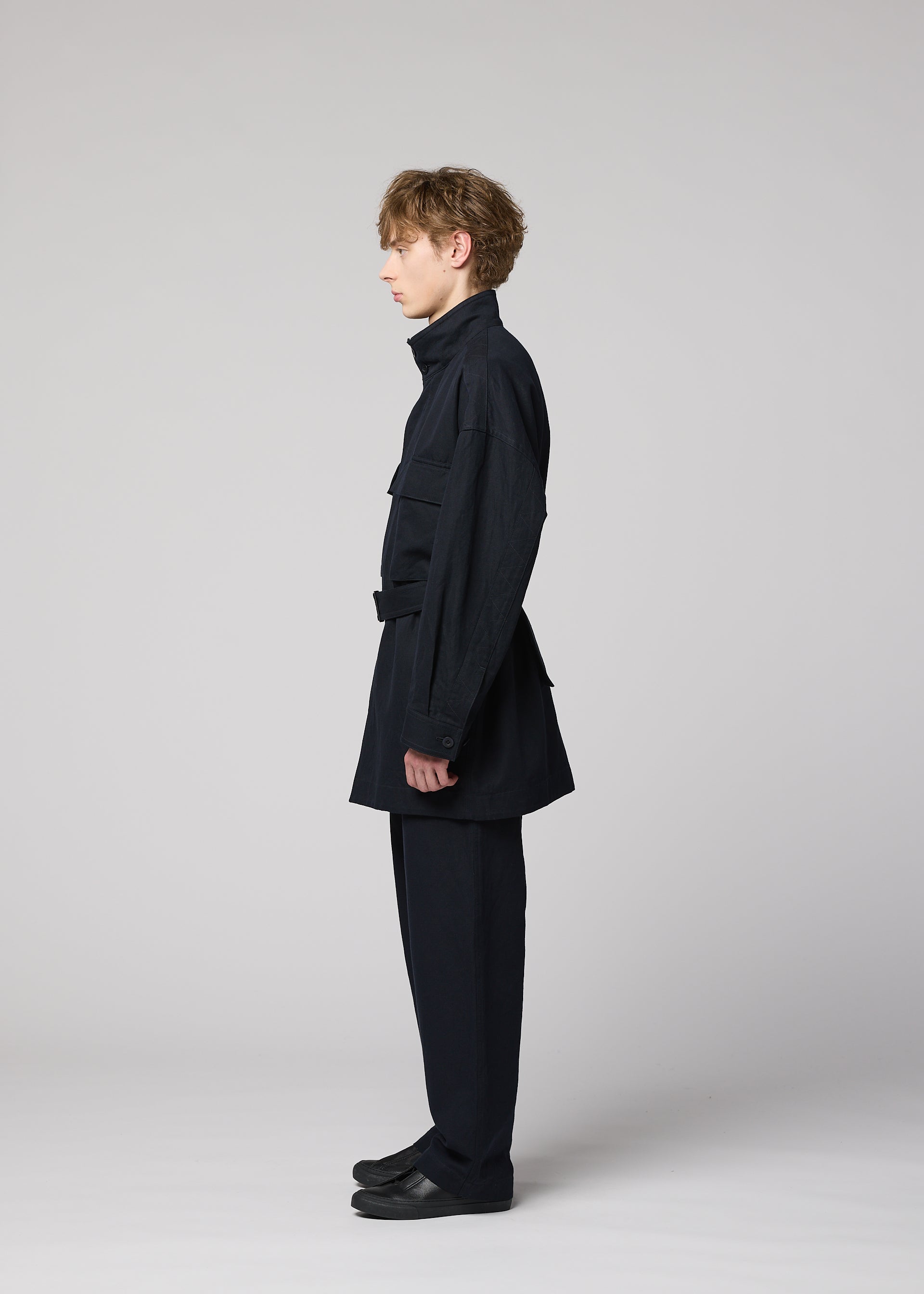 Zoomed view A person stands in profile against a plain gray background, dressed in the IM MEN TOUGH WOOL COTTON jacket and matching black pants, paired with black shoes. The sleek ensemble features luxurious Bishu wool blend fabric, and the individual’s posture is upright, facing left.