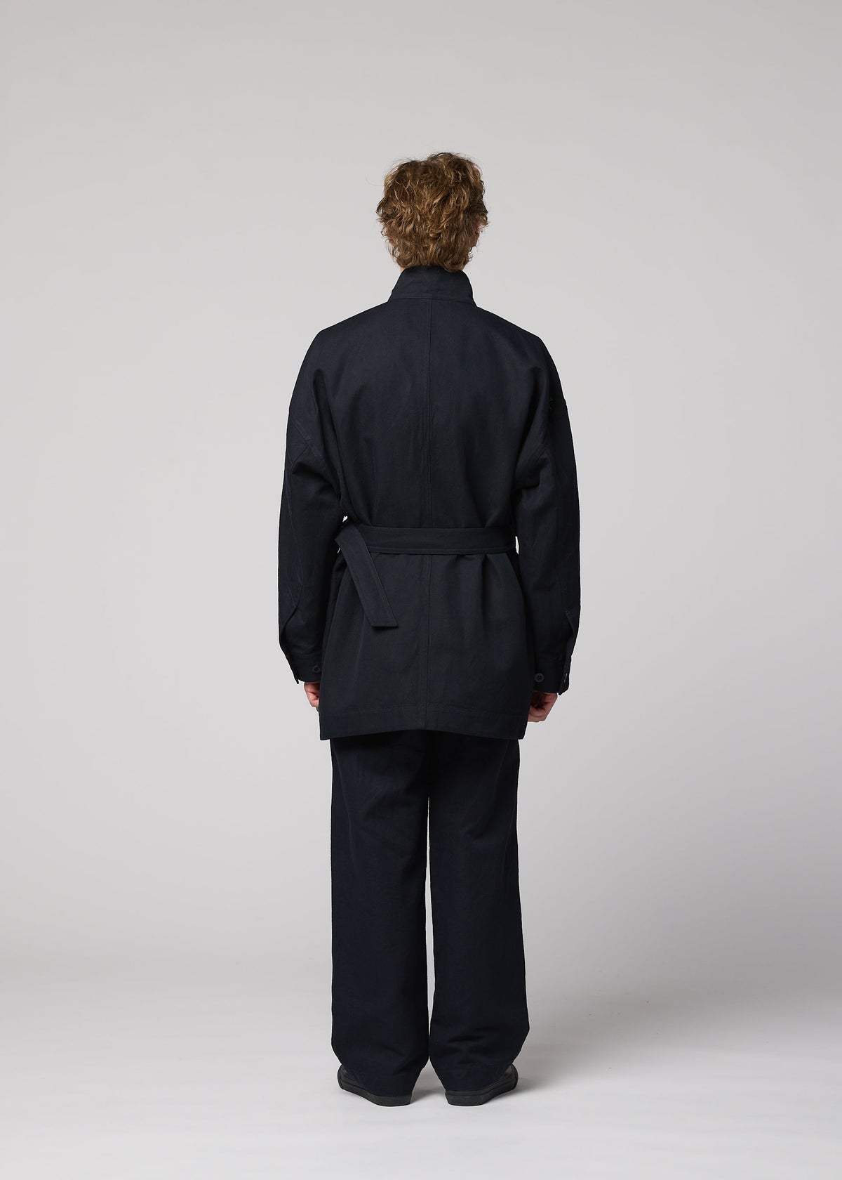 A person with short curly hair stands facing away, wearing a dark TOUGH WOOL COTTON jacket and matching pants by IM MEN, set against a plain light gray background. view 8 of 9