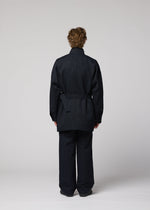 A person with short curly hair stands facing away, wearing a dark TOUGH WOOL COTTON jacket and matching pants by IM MEN, set against a plain light gray background.