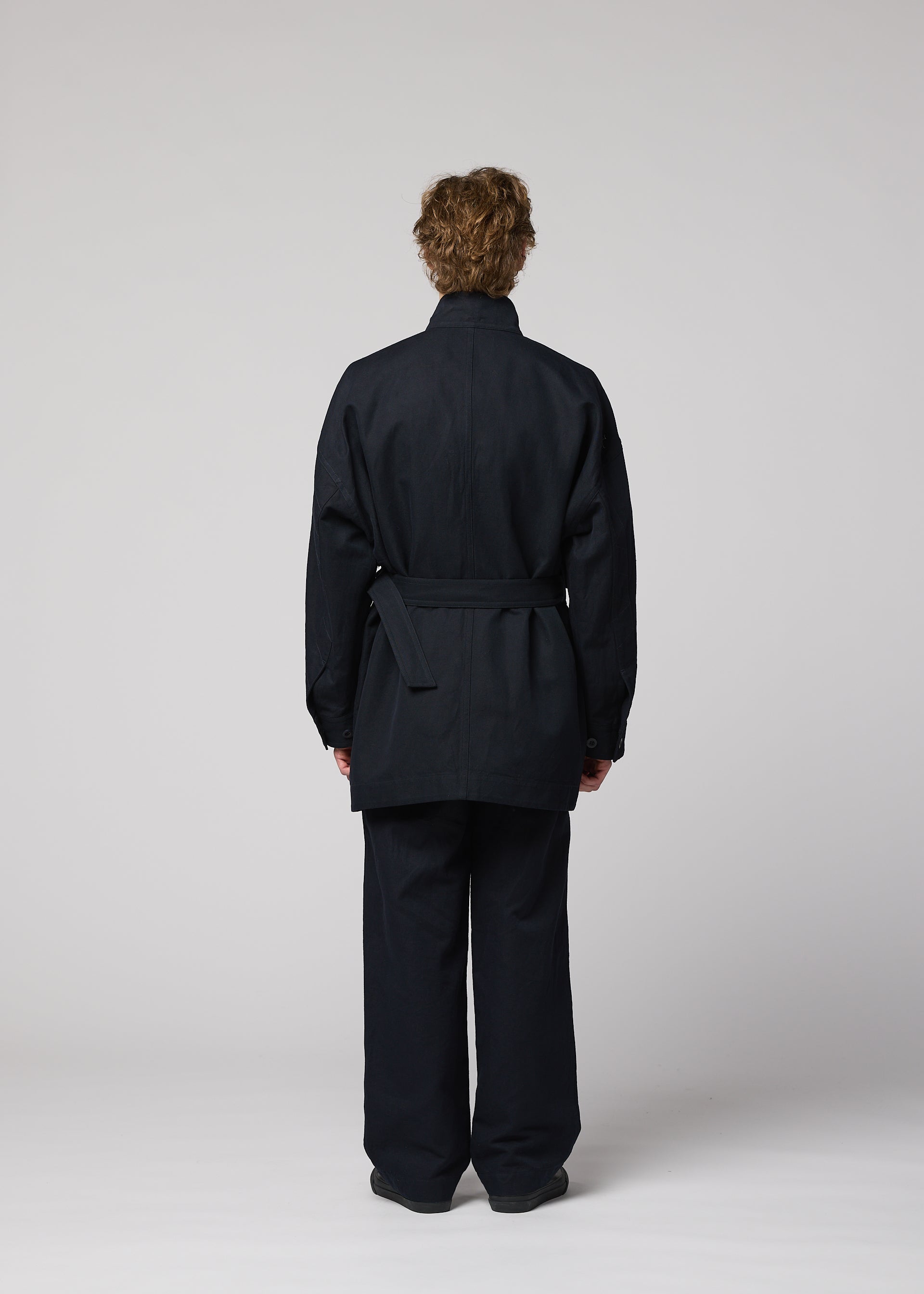 Zoomed view A person with short curly hair stands facing away, wearing a dark TOUGH WOOL COTTON jacket and matching pants by IM MEN, set against a plain light gray background.