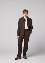 A young person with short, light brown hair stands against a gray background, wearing the IM MEN SUAVE brown wool blend jacket and matching pants, paired with a white shirt. One hand is in their pocket.