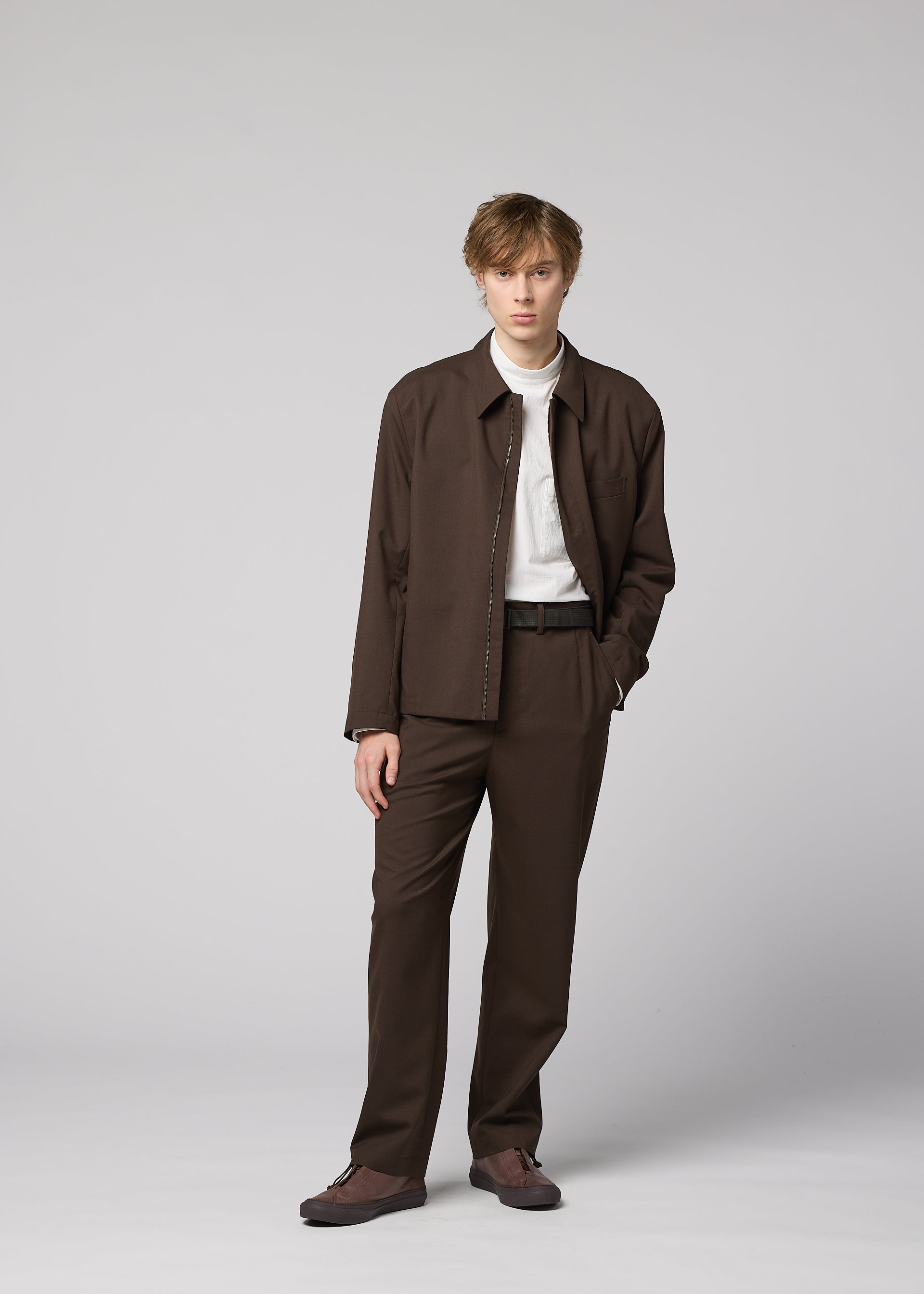 Zoomed view A young person with short, light brown hair stands against a gray background, wearing the IM MEN SUAVE brown wool blend jacket and matching pants, paired with a white shirt. One hand is in their pocket.