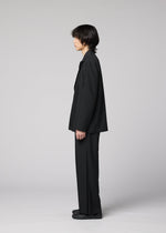 A person with medium-length dark hair stands in profile, wearing a loose jacket, black shoes, and IM MEN’s WINGS BLACK, against a plain light gray background.