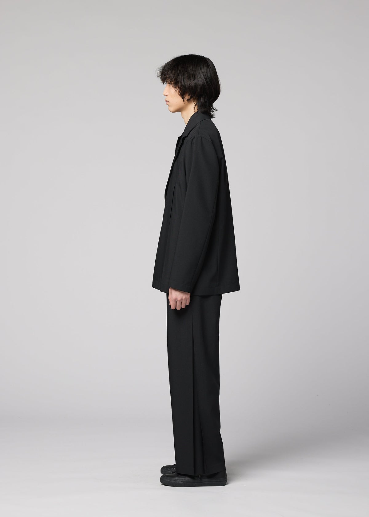 Zoomed view A person with medium-length dark hair stands in profile, wearing a loose jacket, black shoes, and IM MEN’s WINGS BLACK, against a plain light gray background.