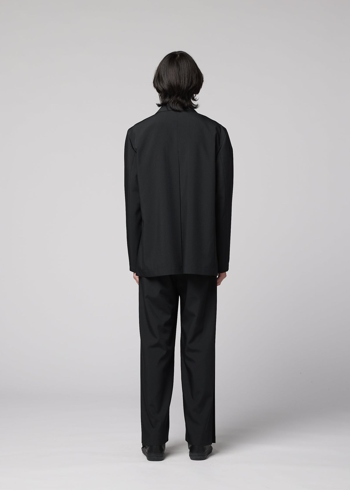 Zoomed view A person with medium-length dark hair, wearing IM MEN’s WINGS BLACK wrinkle resistant jacket and black shoes, stands facing away from the camera against a plain light gray background.