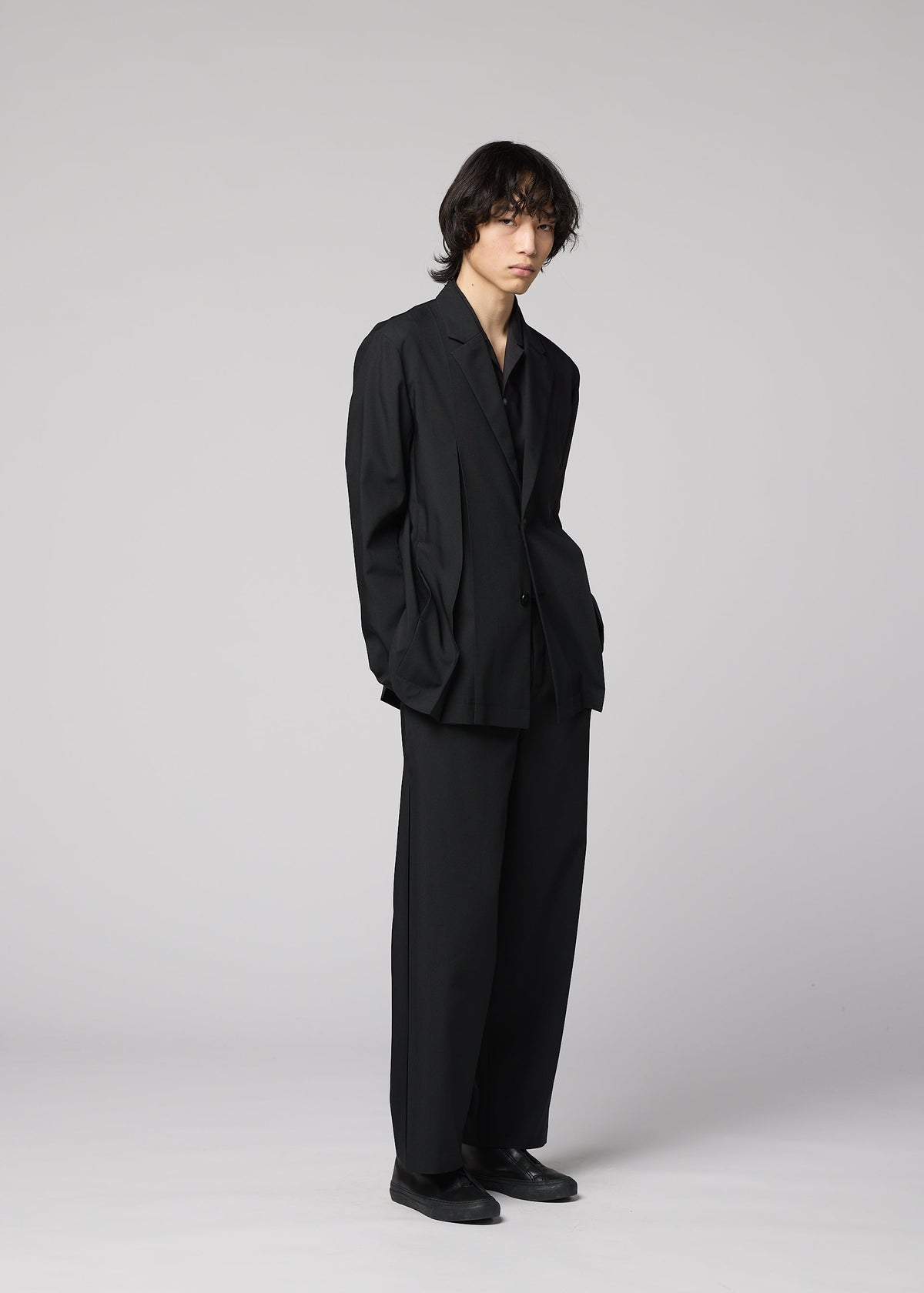 Zoomed view A person with medium-length dark hair stands against a light gray background, wearing IM MEN’s WINGS BLACK jacket with matching trousers, a black shirt, and black shoes.