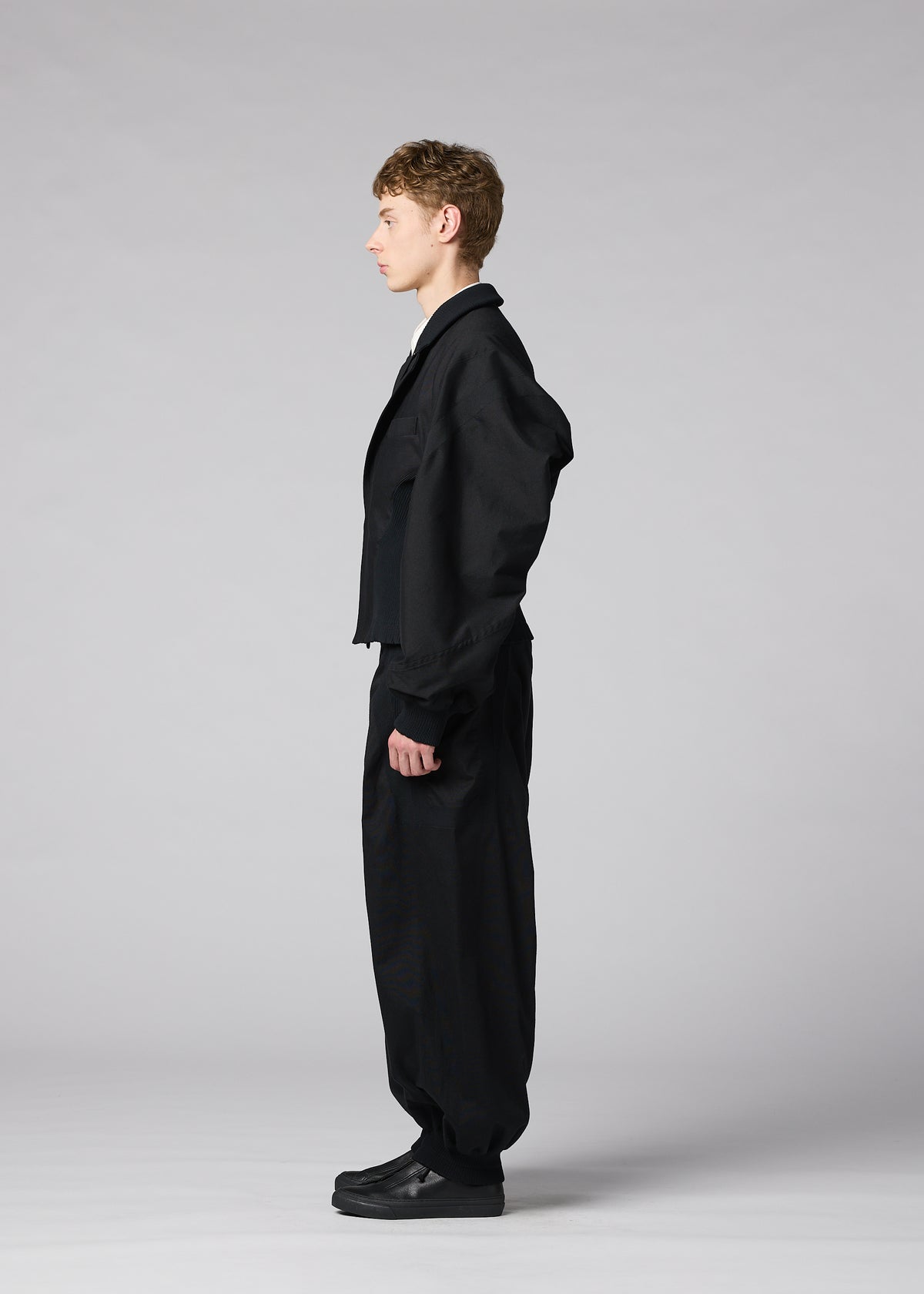 A person stands in profile against a plain gray background, wearing IM MEN’s CLAY—a loose black suit with wide pants and black shoes. The sculptural form of the jacket adds an architectural element, elevating the minimalist ensemble. view 6 of 8