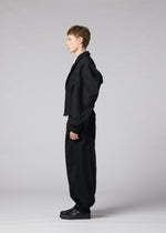 A person stands in profile against a plain gray background, wearing IM MEN’s CLAY—a loose black suit with wide pants and black shoes. The sculptural form of the jacket adds an architectural element, elevating the minimalist ensemble.