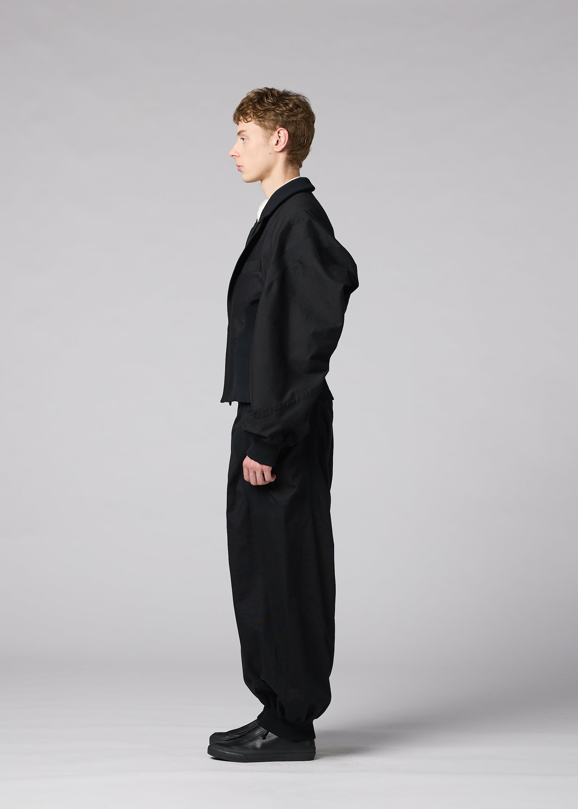 Zoomed view A person stands in profile against a plain gray background, wearing IM MEN’s CLAY—a loose black suit with wide pants and black shoes. The sculptural form of the jacket adds an architectural element, elevating the minimalist ensemble.