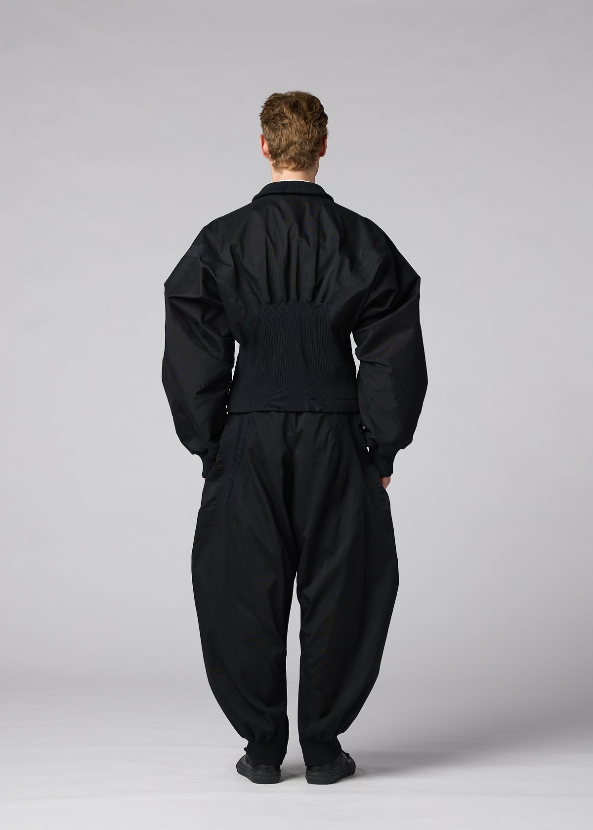 A person stands facing away against a plain, light gray background, wearing IM MEN’s CLAY set: a loose-fitting, all-black ensemble with wide sleeves and pants made from stretch fabric. view 7 of 8
