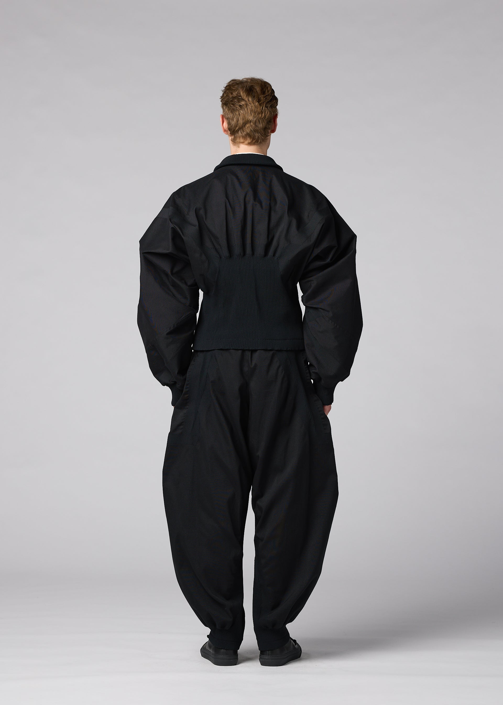 Zoomed view A person stands facing away against a plain, light gray background, wearing IM MEN’s CLAY set: a loose-fitting, all-black ensemble with wide sleeves and pants made from stretch fabric.