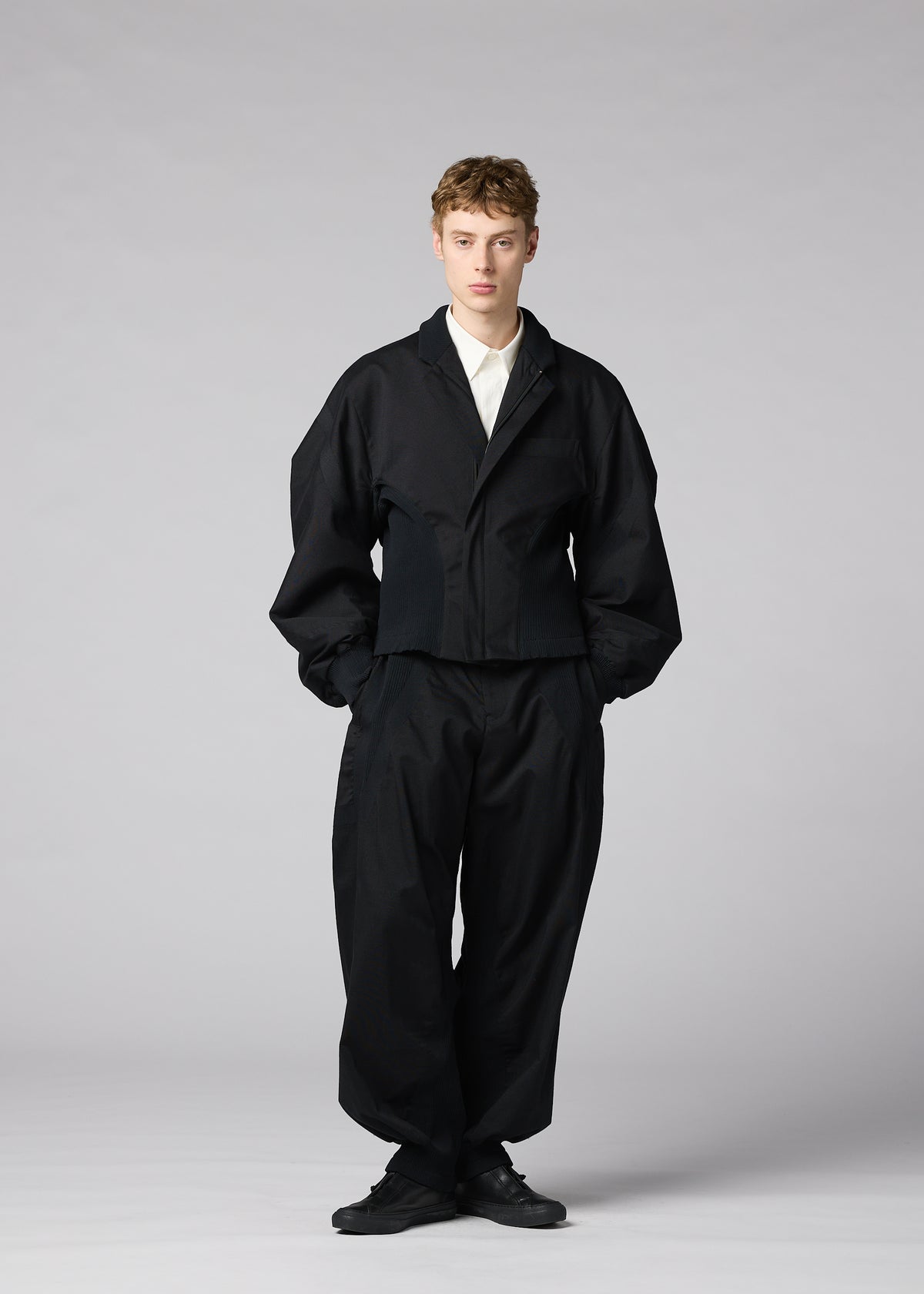 A person stands facing forward against a plain light gray background, hands in pockets and expression neutral, wearing the IM MEN “CLAY” jacket—a loose-fitting black sculptural form—paired with a white shirt and black shoes. view 8 of 8
