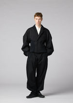 A person stands facing forward against a plain light gray background, hands in pockets and expression neutral, wearing the IM MEN “CLAY” jacket—a loose-fitting black sculptural form—paired with a white shirt and black shoes.
