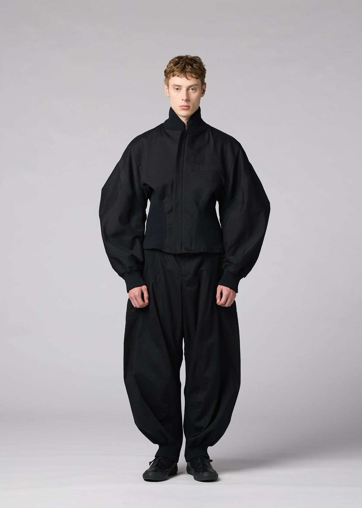 A person stands facing forward, wearing the IM MEN “CLAY” jacket featuring a sculptural form with a high neck and voluminous sleeves, paired with loose black pants. The background is plain and light gray. view 5 of 8