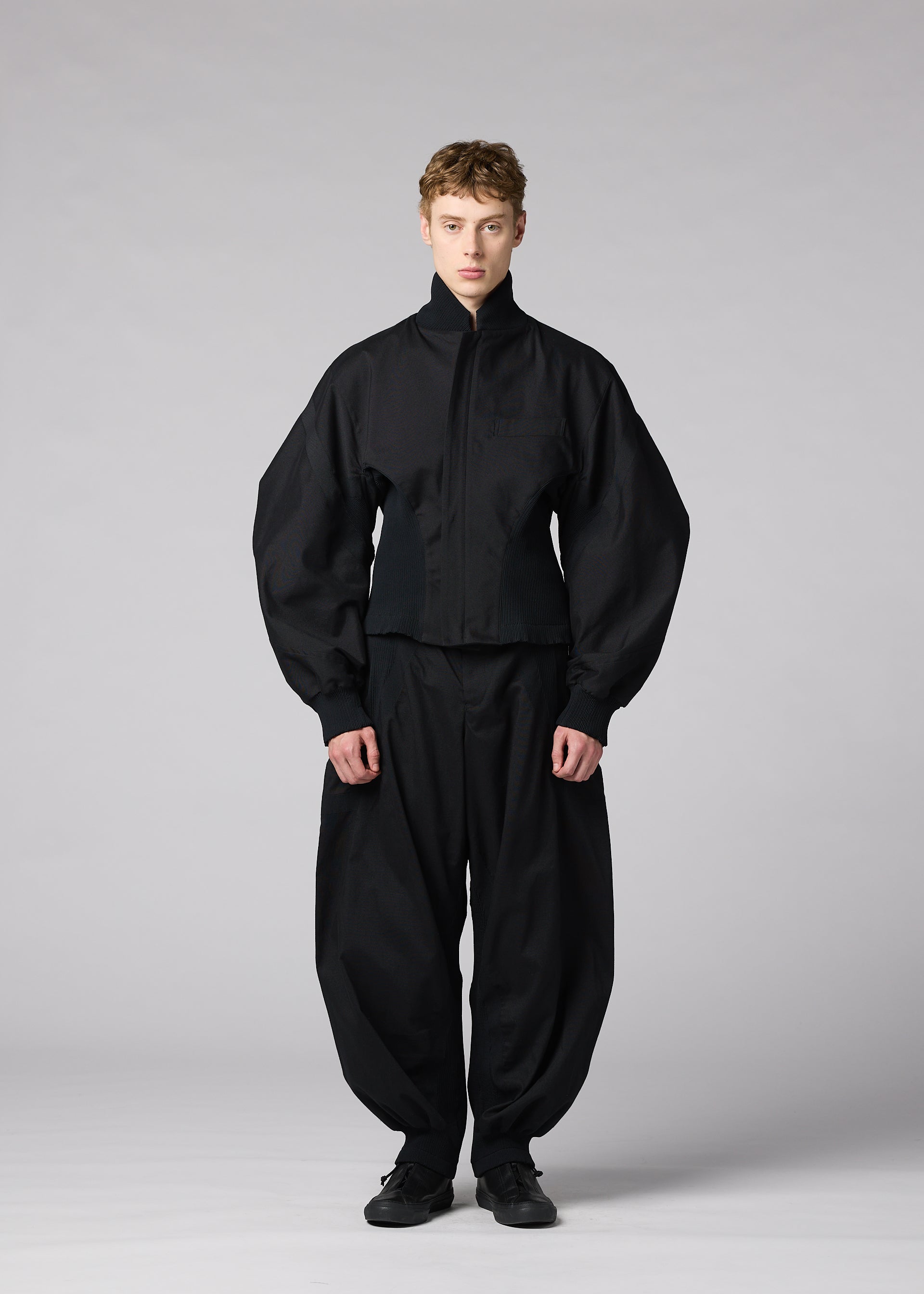 Zoomed view A person stands facing forward, wearing the IM MEN “CLAY” jacket featuring a sculptural form with a high neck and voluminous sleeves, paired with loose black pants. The background is plain and light gray.