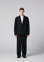A person stands facing forward against a plain light gray background, wearing IM MEN’s CLAY—a loose-fitting black suit with a sculptural silhouette—paired with a white collared shirt and black shoes. Their hands are at their sides and their expression is neutral.