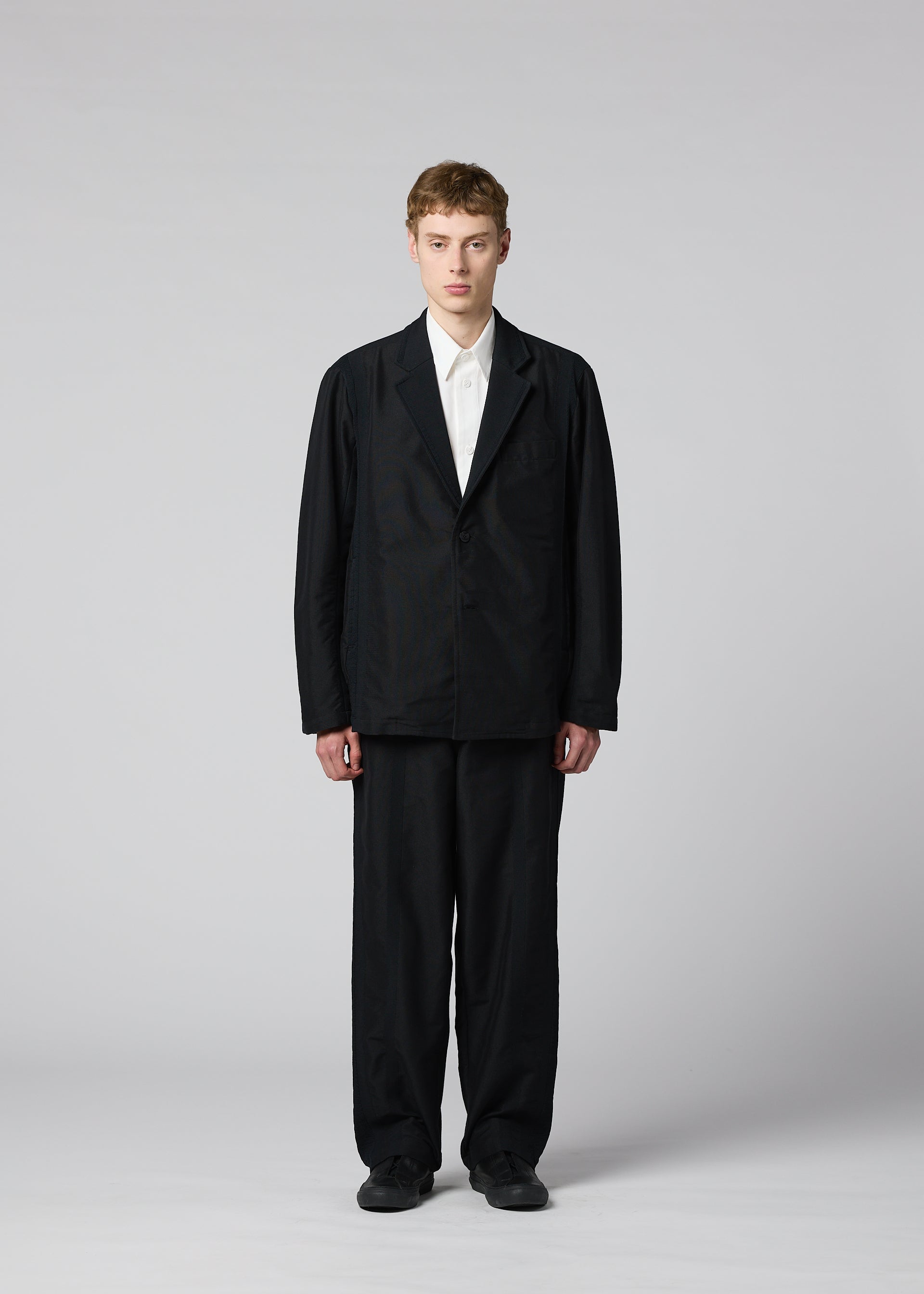 Zoomed view A person stands facing forward against a plain light gray background, wearing IM MEN’s CLAY—a loose-fitting black suit with a sculptural silhouette—paired with a white collared shirt and black shoes. Their hands are at their sides and their expression is neutral.