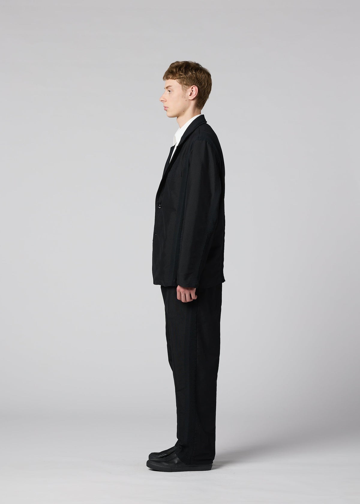 A man wearing the IM MEN “CLAY” black sculptural suit with matching black shoes stands in profile against a plain light gray background, facing left, his arms relaxed at his sides. view 6 of 8