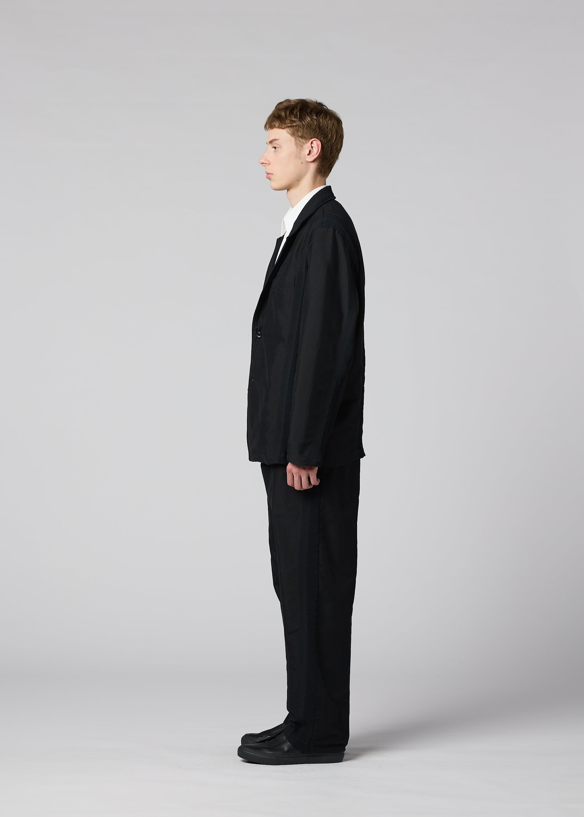 Zoomed view A man wearing the IM MEN “CLAY” black sculptural suit with matching black shoes stands in profile against a plain light gray background, facing left, his arms relaxed at his sides.