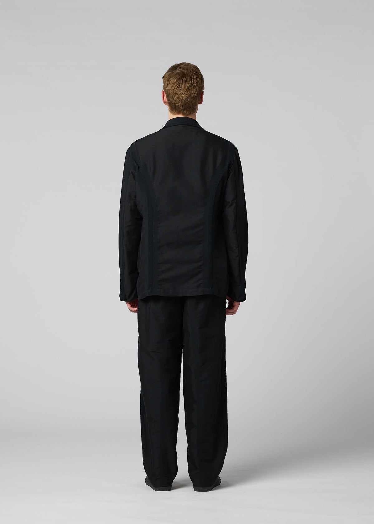 A person with short light brown hair stands facing away, wearing the loose-fitting black CLAY textile jacket and matching black pants by IM MEN against a plain light gray background. view 7 of 8