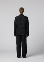 A person with short light brown hair stands facing away, wearing the loose-fitting black CLAY textile jacket and matching black pants by IM MEN against a plain light gray background.
