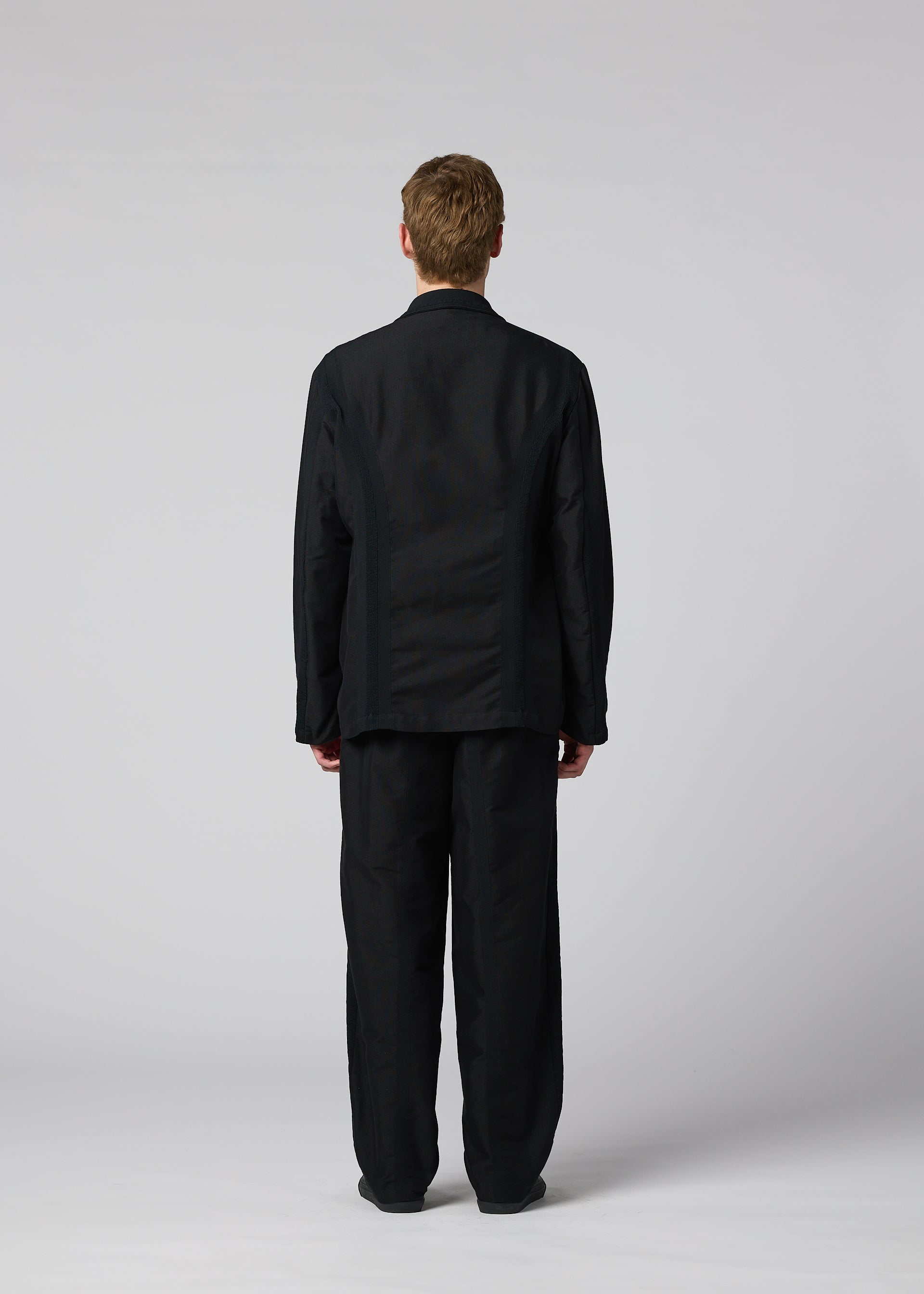 Zoomed view A person with short light brown hair stands facing away, wearing the loose-fitting black CLAY textile jacket and matching black pants by IM MEN against a plain light gray background.