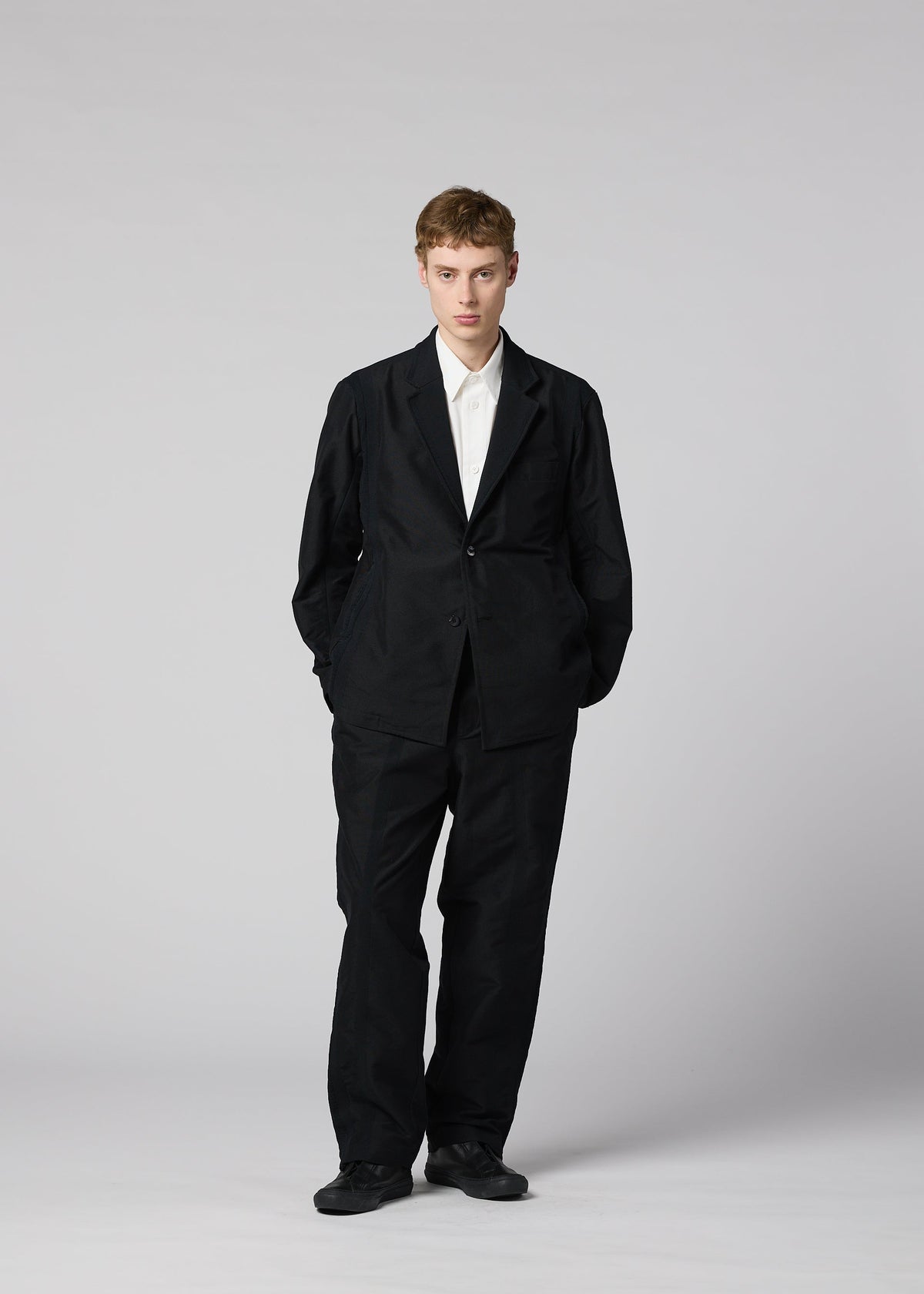 A person stands against a plain, light gray background, wearing IM MEN’s CLAY—a sculptural black suit jacket paired with a crisp white shirt and black shoes. The distinctive form of the CLAY jacket adds subtle dimension to the ensemble. With hands in their pockets, they face the camera directly. view 8 of 8