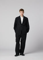 A person stands against a plain, light gray background, wearing IM MEN’s CLAY—a sculptural black suit jacket paired with a crisp white shirt and black shoes. The distinctive form of the CLAY jacket adds subtle dimension to the ensemble. With hands in their pockets, they face the camera directly.