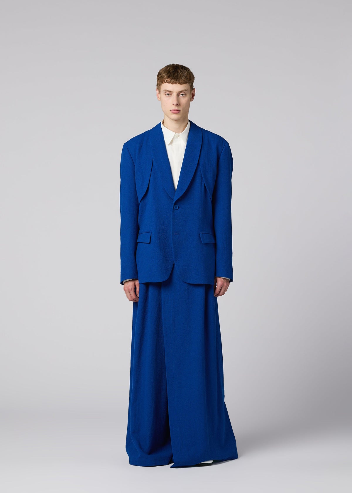 Zoomed view A person stands facing forward in an IM MEN SHRINK WOOL oversized royal blue jacket and wide-legged trousers over a white shirt, against a plain light grey background.