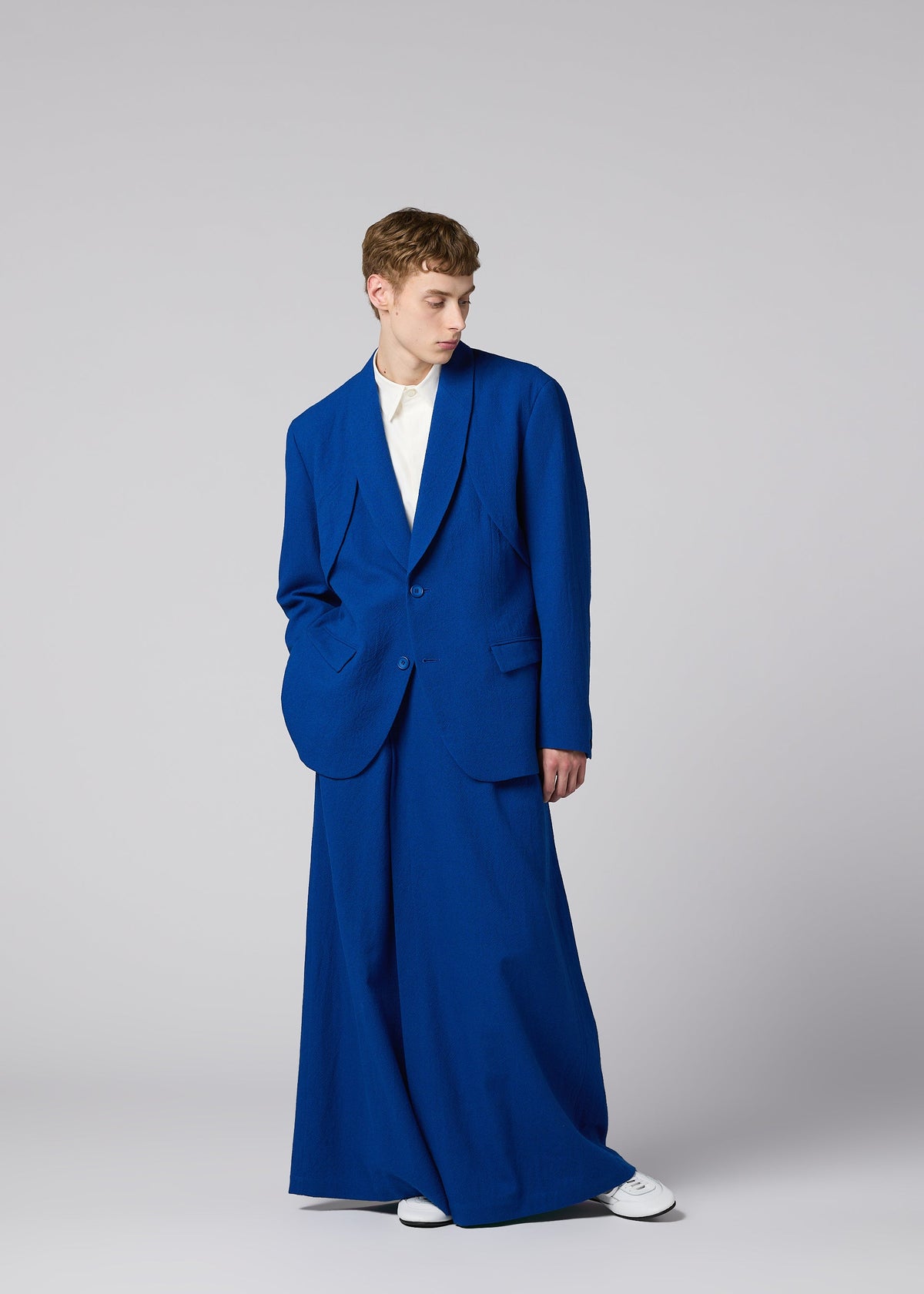 Zoomed view A person stands against a gray background wearing SHRINK WOOL by IM MEN: an oversized royal blue hard-twist wool jacket, wide-legged pants, white shirt, and white shoes. Autumn Winter 2026/27.