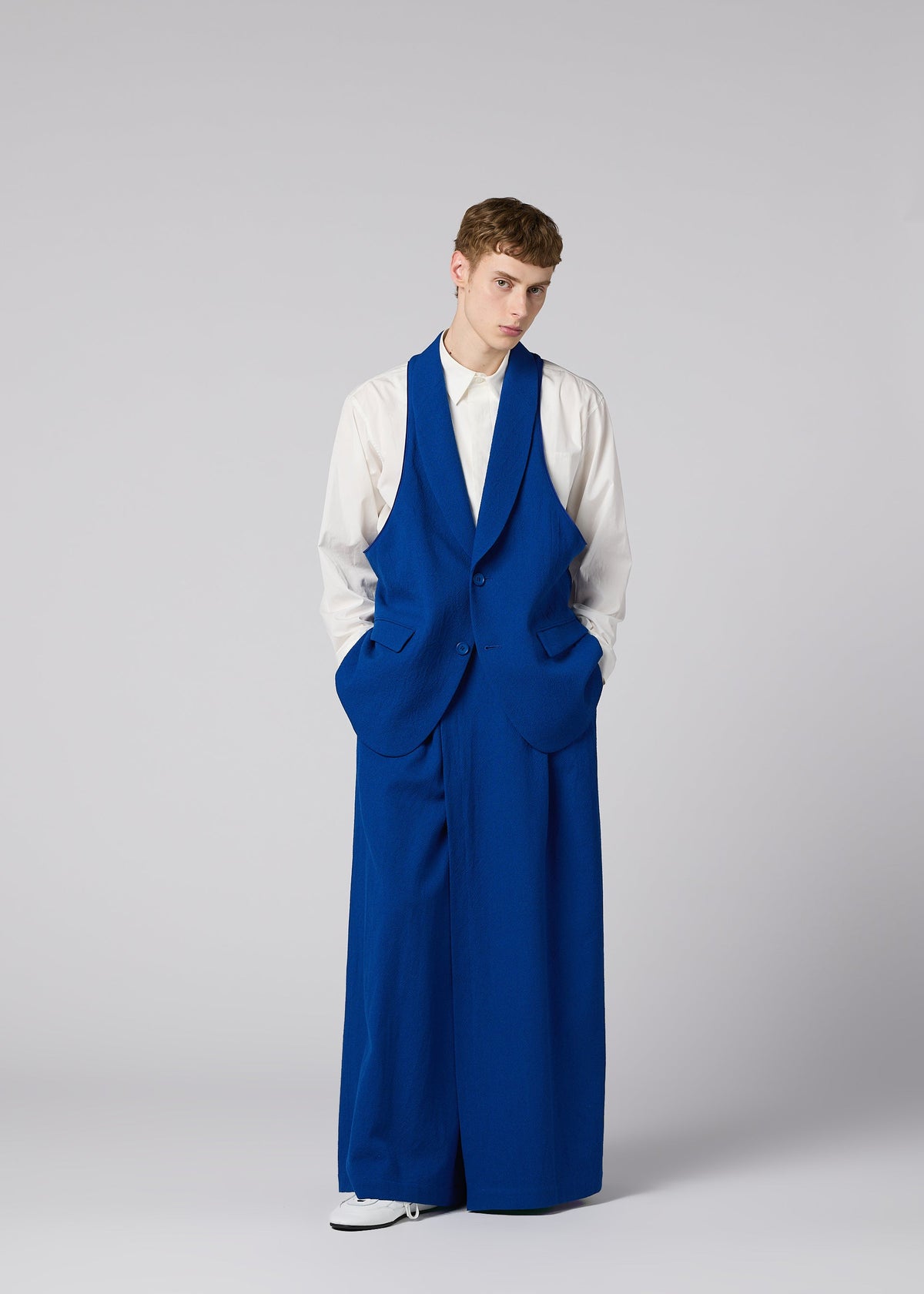 A person stands against a plain background wearing a white shirt and bright blue, oversized SHRINK WOOL jacket by IM MEN, with matching wide-leg pants and hands in pockets. view 14 of 17