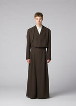 A person wears the IM MEN SHRINK WOOL suit—a brown, long, wide-legged set with a collarless jacket in hard-twist wool over a white top—standing against a plain background, looking straight ahead neutrally.