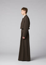 A person in profile poses against a light gray background wearing the IM MEN SHRINK WOOL brown collarless jacket and matching wide-legged pants, both in hard-twist wool with a crepe look.