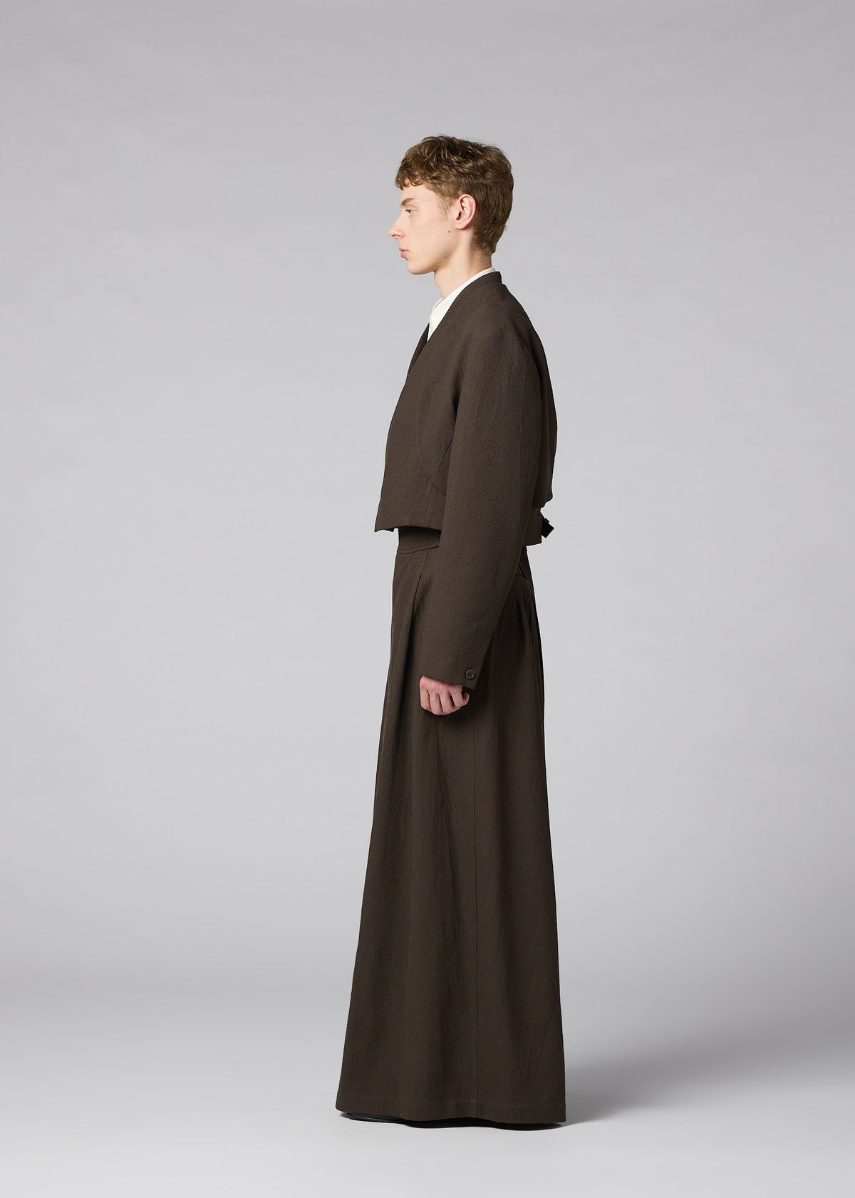 Zoomed view A person in profile poses against a light gray background wearing the IM MEN SHRINK WOOL brown collarless jacket and matching wide-legged pants, both in hard-twist wool with a crepe look.